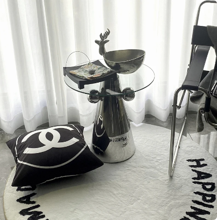 A modern designer side table with a glass top and stainless steel legs, placed on a white rug with a black and white pillow, next to a chair in a bright room.