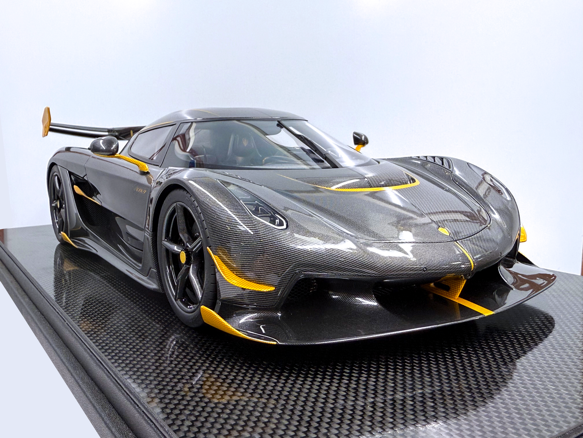 Vinklad sidovy av Koenigsegg Jesko – Carbon Fury Edition i skala 1:8 som visar exponerad kolfiberkaross, gula linjedetaljer och aggressiv aerodynamik från front till bak.